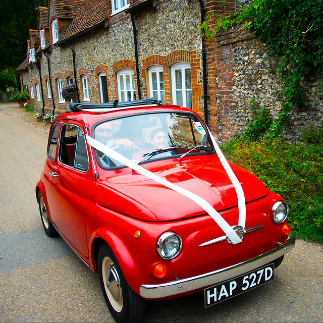 Image of a man driving a small red car with wedding ribbons on. Image of a man and womans hands during a wedding
